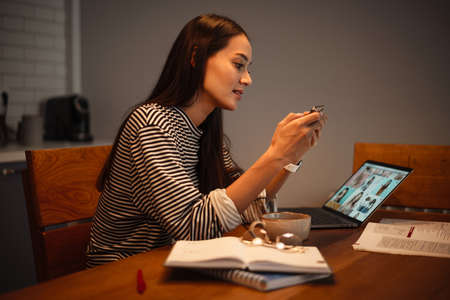 Focused charming woman using cellphone while working with laptop at home kitchen