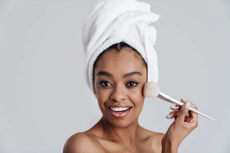 Happy african american woman in bath towel using powder brush isolated over white backgroundの写真素材
