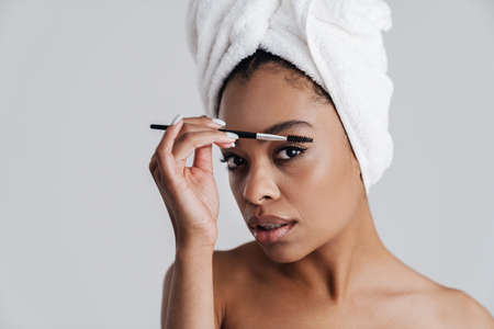 Focused african american woman in bath towel using eyebrow brush isolated over white backgroundの写真素材