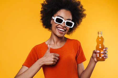 Joyful african american girl in sunglasses drinking soda and showing ok sign isolated over yellow backgroundの写真素材