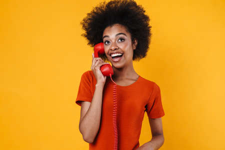 Cheerful african american girl smiling and talking on retro phone isolated over yellow backgroundの写真素材