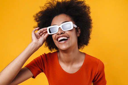 Joyful african american girl in sunglasses laughing at camera isolated over yellow backgroundの写真素材
