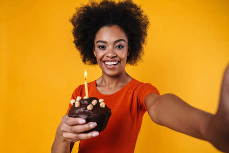 Joyful african american girl taking selfie while holding cake isolated over yellow backgroundの写真素材