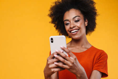 Joyful african american girl smiling while using mobile phone isolated over yellow background