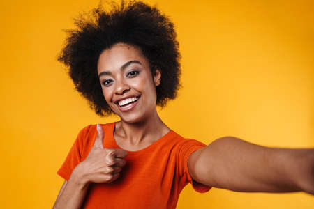 Joyful african american girl taking selfie while showing thumb up isolated over yellow backgroundの写真素材