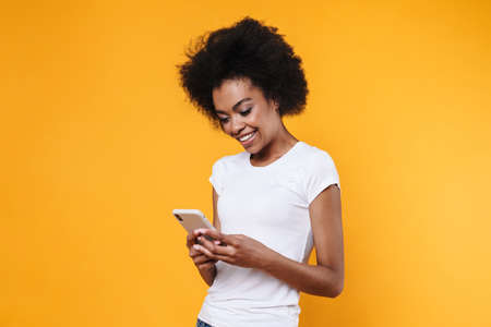 Happy young african american woman smiling and holding cellphone isolated over yellow background