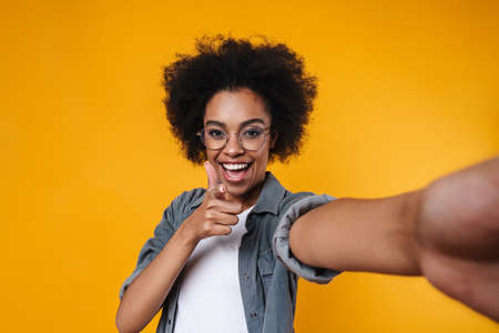 Joyful african american girl showing thumb up while taking selfie photo isolated over yellow...