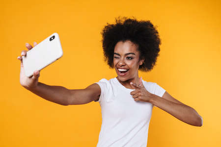 Happy young african american woman pointing finger at cellphone while taking selfie photo isolated over yellow backgroundの写真素材