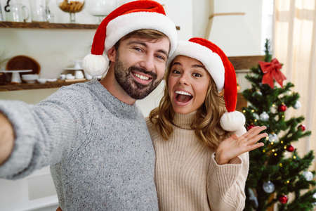 Beautiful excited couple in santa claus hats hugging and taking selfie photo in cozy kitchenの写真素材