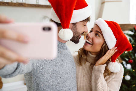 Image of young couple man and woman wearing christmas outfit standing in bright kitchen and taking selfie photo on mobile phoneの写真素材