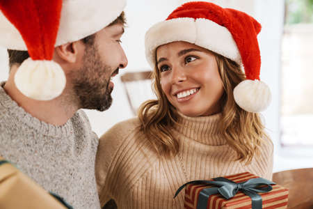 Romantic happy couple in santa claus hats giving gifts at each other while hugging in cozy roomの写真素材