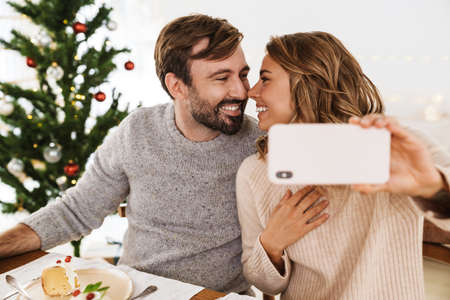 Charming cheerful couple taking selfie photo on cellphone while having Christmas dinner in cozy roomの写真素材