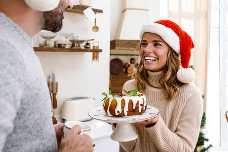 Happy woman in santa claus hat showing Christmas pie to her boyfriend in cozy kitchenの写真素材