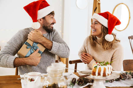Happy woman in santa claus hat giving gift box to his boyfriend while having Christmas dinner in cozy roomの写真素材