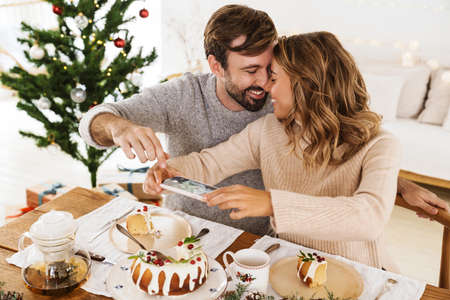Charming cheerful couple taking photo on cellphone while having Christmas dinner in cozy roomの写真素材