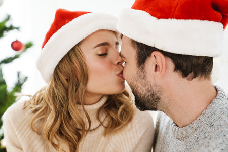 Beautiful happy couple in santa claus hats smiling and kissing indoorsの写真素材