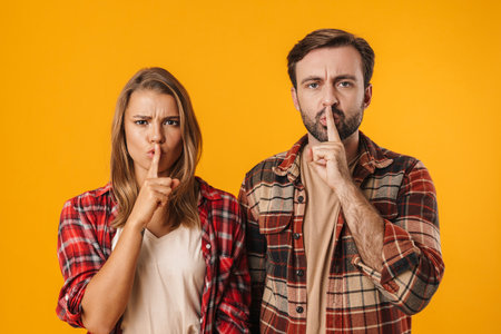 Caucasian displeased couple showing silence gesture at camera isolated over yellow background