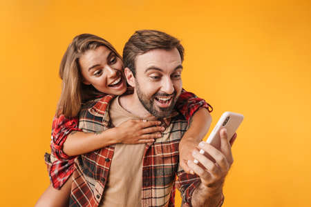 Excited man using cellphone while piggyback riding his smiling girlfriend isolated over yellow backgroundの写真素材