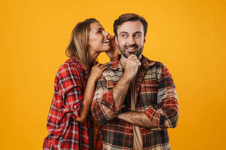 Pleased blonde woman whispering secret at smiling man isolated over yellow backgroundの写真素材