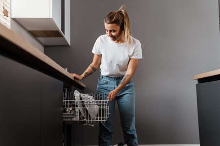 Beautiful happy girl smiling while using dishwasher at home kitchenの写真素材