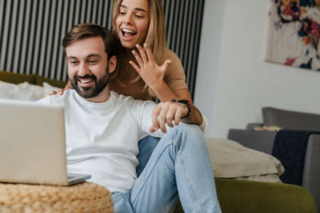 Happy young attractive couple on a video call with their friends via laptop computer, sitting in the bedroom, showing engagement ringの写真素材