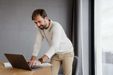 Handsome smiling man in earphones working with laptop while standing indoors