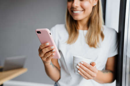 Beautiful smiling girl using mobile phone and drinking coffee indoorsの写真素材
