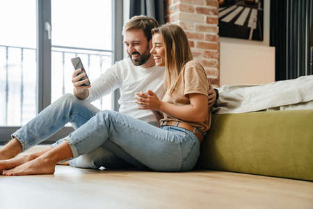Happy beautiful couple using mobile phone while relaxing at homeの写真素材