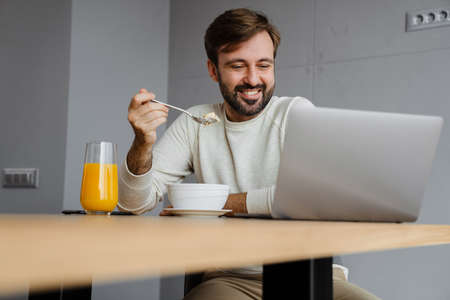 Handsome happy man using laptop while having breakfast at home kitchenの写真素材