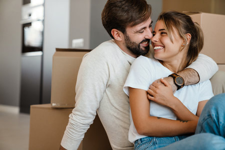 Beautiful couple smiling and hugging while sitting on floor with cardboard boxes in new apartmentの写真素材
