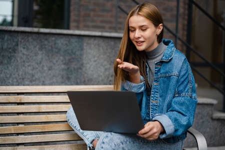 Beautiful happy student girl blowing air kiss and using laptop while sitting on bench at city streetの写真素材