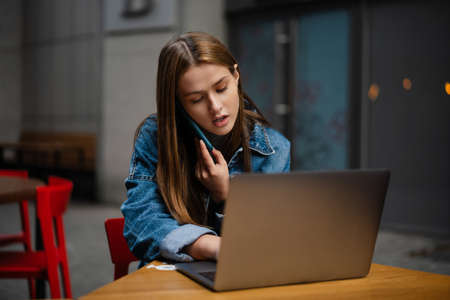 Beautiful focused student girl talking on cellphone while working with laptop in street cafe outdoorsの写真素材