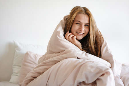 Beautiful cheerful nice girl covered in blanket smiling and sitting on bed at homeの写真素材