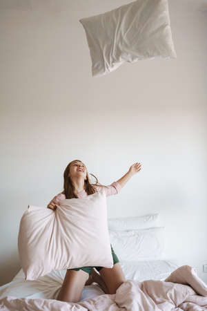 Beautiful excited nice girl laughing and making fun with pillows on bed at home
