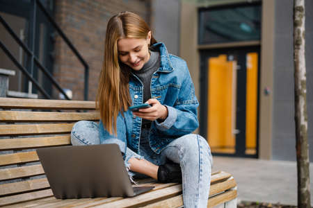 Beautiful happy student girl using cellphone and laptop while sitting on bench at city streetの写真素材