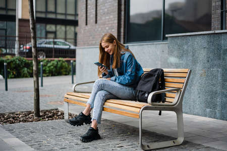Beautiful young woman wearing denim clothes sitting on a street bench, using mobile phoneの写真素材