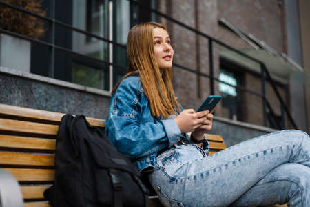 Beautiful pleased student girl using cellphone while sitting on bench at city streetの写真素材