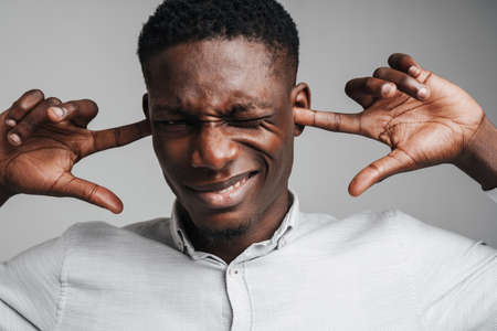 Handsome smiling african man in white shirt covering his ears, headache from loud noise, isolated on...