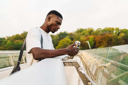 Handsome african sportsman using mobile phone, holding water bottle at the city bridge
