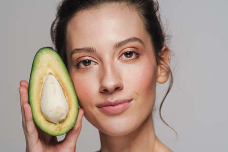Pleased half-naked woman looking at camera while posing with avocado isolated over grey backgroundの写真素材