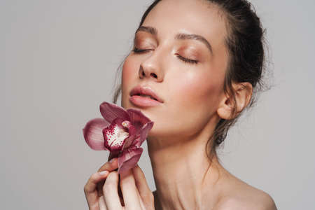 Sensual half-naked woman posing with orchid on camera isolated over grey background