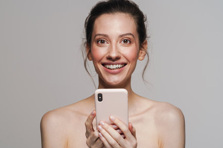 Happy half-naked woman smiling while using mobile phone isolated over grey backgroundの写真素材