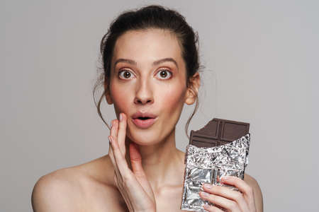 Surprised half-naked woman posing with chocolate on camera isolated over grey backgroundの写真素材