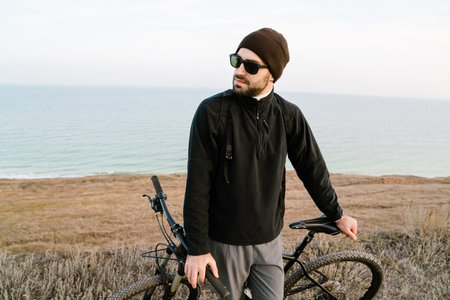 Handsome unshaven guy in sunglasses standing with his bicycle on coast