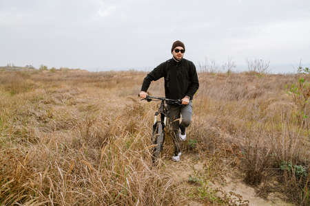 Athletic unshaven guy in sunglasses walking with his bicycle on fieldの写真素材