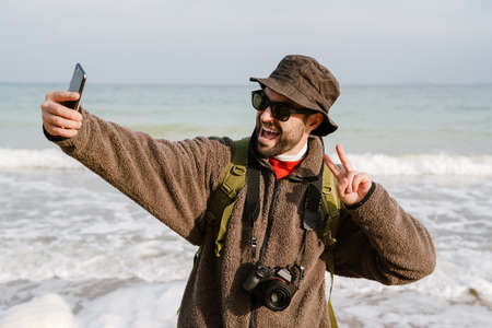 Happy photographer showing peace sign while taking selfie on cellphone at beach