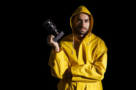 Serious photographer in raincoat posing with digital camera isolated over black background