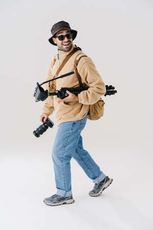 Excited young photographer walking with digital camera and tripod isolated over white background