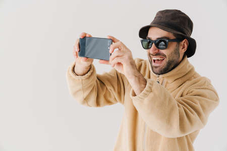 Portrait of a happy young man wearing beige jacket, cap and sunglasses on isolated white background, taking a selfieの写真素材