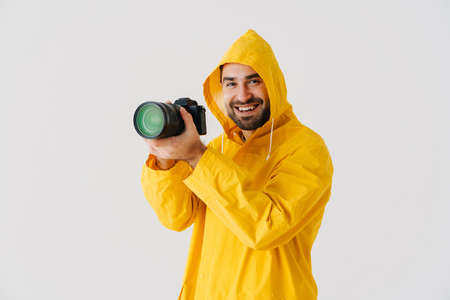 Cheerful photographer in raincoat taking photo on digital camera isolated over white backgroundの写真素材